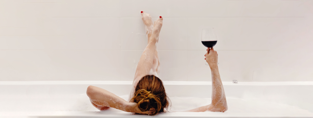 Una mujer relajándose en una bañera con las piernas levantadas y cruzadas, sosteniendo una copa de vino tinto, centrándose en la tranquilidad y el ocio.